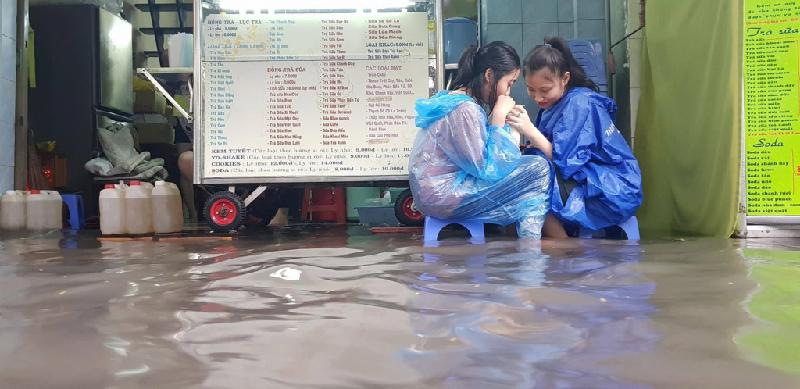 Hai cô gái mặc áo mưa, ngồi uống trà sữa giữa nước ngập lênh láng.