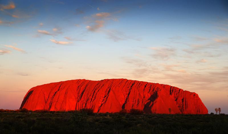 Núi Uluru nhìn từ xa dưới ánh hoàng hôn. Núi Uluru hay Đá Ayers là một ngọn núi hình thành do quá trình bồi đắp của cát giữa hoang mạc miền trung Australia. Uluru nằm trong lãnh thổ của người bản địa Anangu, thuộc khu vực Công viên quốc gia Uluru-Kata Tjuta. Nơi đây có nhiều mạch nước, hang đá và tranh vẽ cổ. Uluru được UNESCO công nhận là di sản thế giới. Ảnh: Getty.